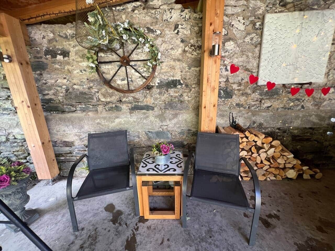 A rustic room with a stone wall, a wooden table with a flower vase, two black chairs, and a wheel with greenery.