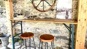 Una pared de piedra rústica con techo de madera, un adorno de rueda metálica y dos taburetes.