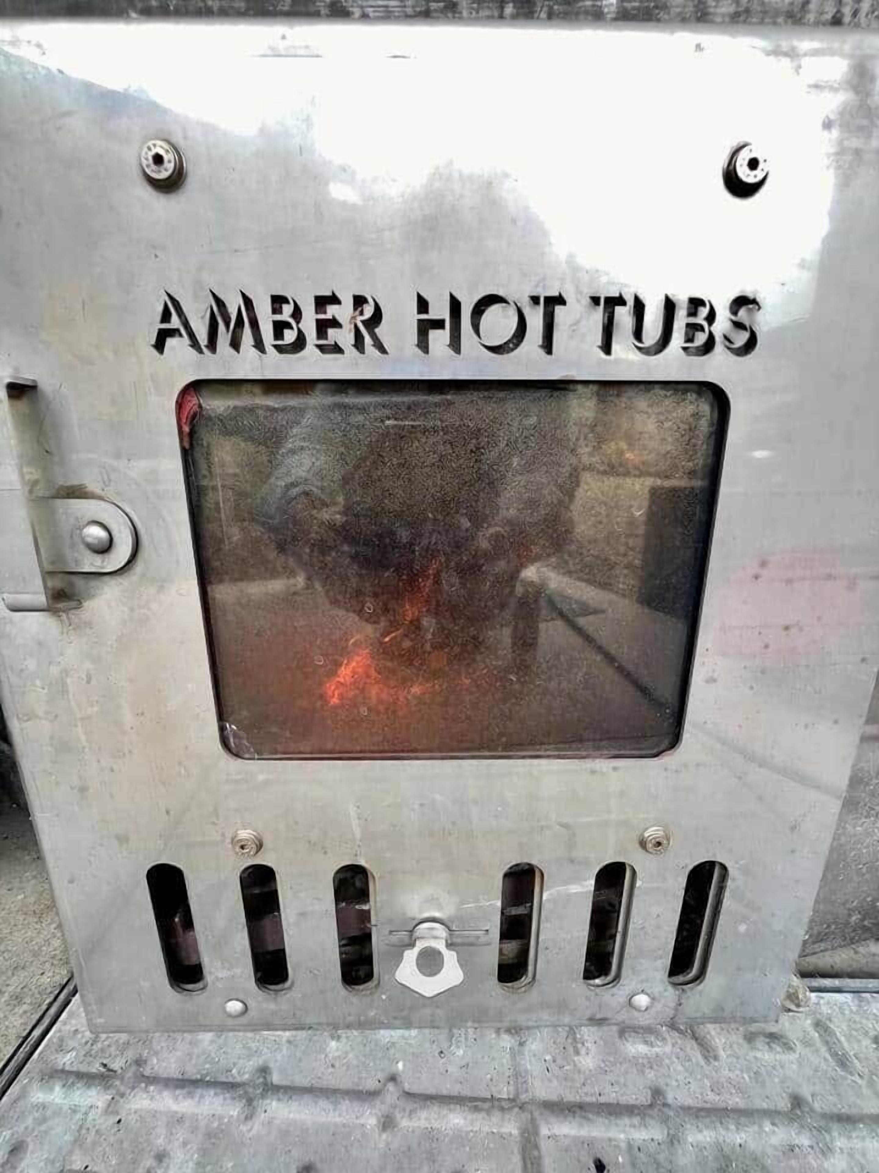 Eine Metallkonstruktion mit dem Schriftzug 'AMBER HOT TUBS' und einem Fenster, das ein Feuer reflektiert.
