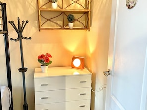Eine weiße Kommode mit Schubladen, ein kleiner Topf mit roten Blüten, ein an der Wand befestigtes Regal mit zwei Topfpflanzen und eine Stehlampe.