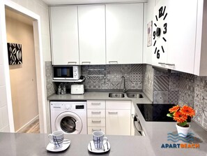 Eine moderne Küche mit weißen Schränken, einer Mikrowelle, einem Toaster, einer Spüle und einer Waschmaschine. Auf der Arbeitsfläche stehen zwei Tassen.