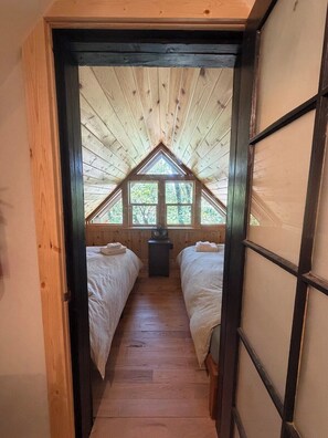 Un pasillo estrecho que conduce a un dormitorio con dos camas, un ventilador y una ventana con vista a árboles.