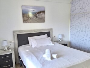 Ein ordentlich bezogenes Bett mit weißen Leinen und einem Handtuch, zwei Nachttische mit Lampen und ein Gemälde mit Strandmotiv an der Wand.