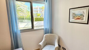 A room with a checkered armchair, a bed with an orange towel, a window with blue curtains, and a framed picture on the wall. - Beautiful beachfront apartment with pool and private parking (Miami Playa)