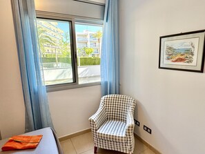 Ein Zimmer mit einem karierten Sessel, einem Bett mit orangefarbenem Handtuch, einem Fenster mit blauen Vorhängen und einem gerahmten Bild an der Wand.