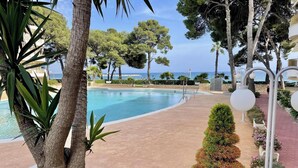 A pool area with a curved edge, surrounded by palm trees and other greenery, with a view of the sea in the background. - Family Seafront Apartment - Pool, Air Conditioning, Terrace (Cambrils)