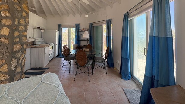 A spacious room with a stone wall, a ceiling fan, a dining area with wicker chairs, and large windows with blue curtains. - On the beach, Private path to the ocean beach. (Cat Island)