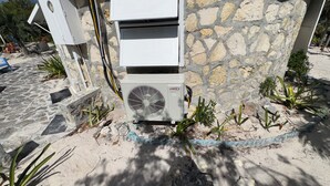 An air conditioning unit mounted on a stone wall, surrounded by small plants and a blue-painted edge. - On the beach, Private path to the ocean beach. (Cat Island)