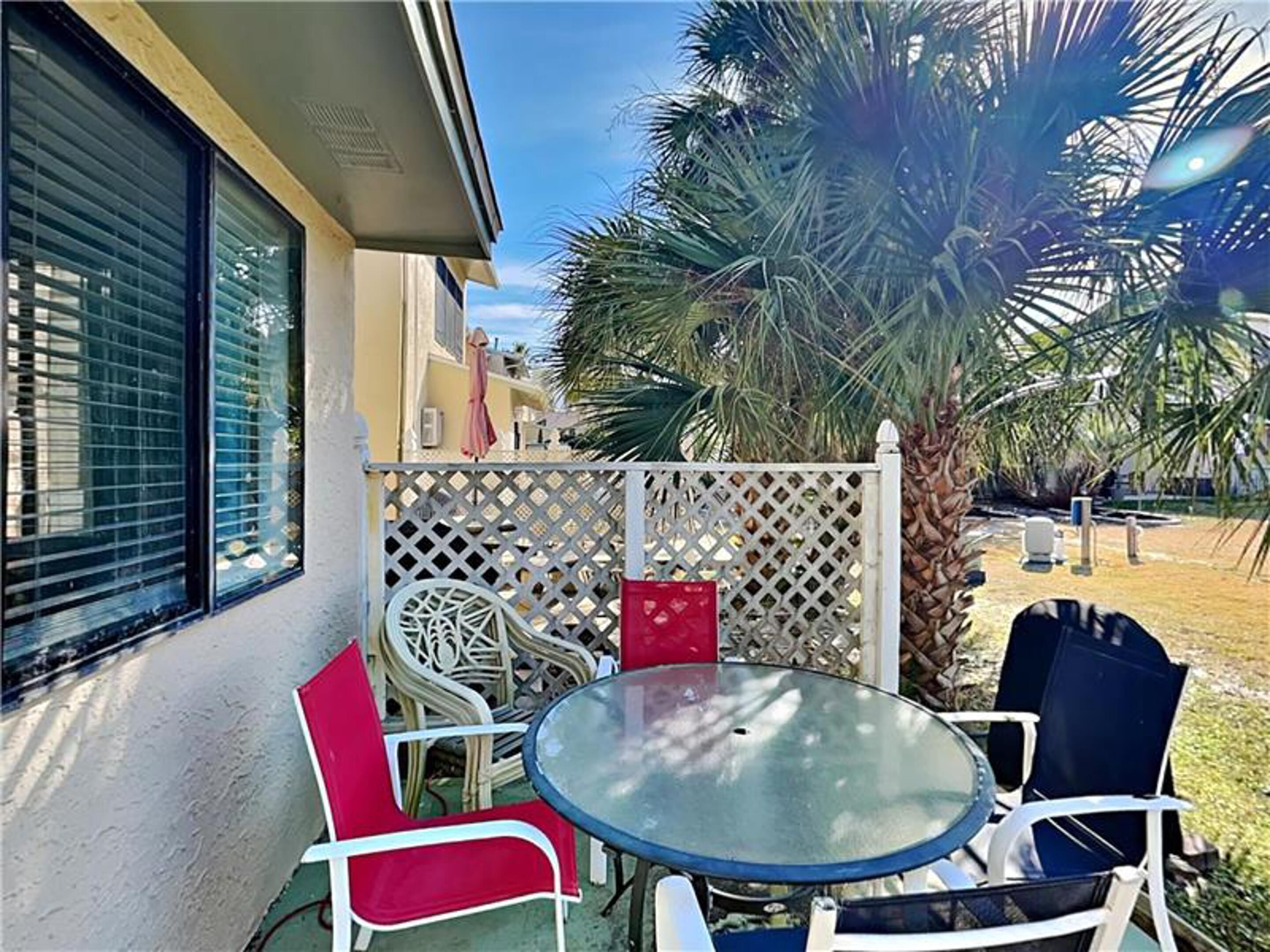 An outdoor patio area with a round glass table and four chairs, a white lattice fence, and a palm tree.