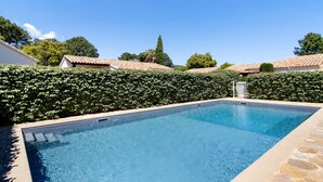 Ein rechteckiger Pool mit Steinrand, umgeben von einer Hecke und einer überdachten, mit Fliesen versehenen Struktur im Hintergrund.