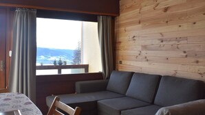 Ein gemütliches Zimmer mit einer Holzwand, einem grauen Sofa, einem Holzstuhl und einem Tisch mit gemustertem Tischläufer.