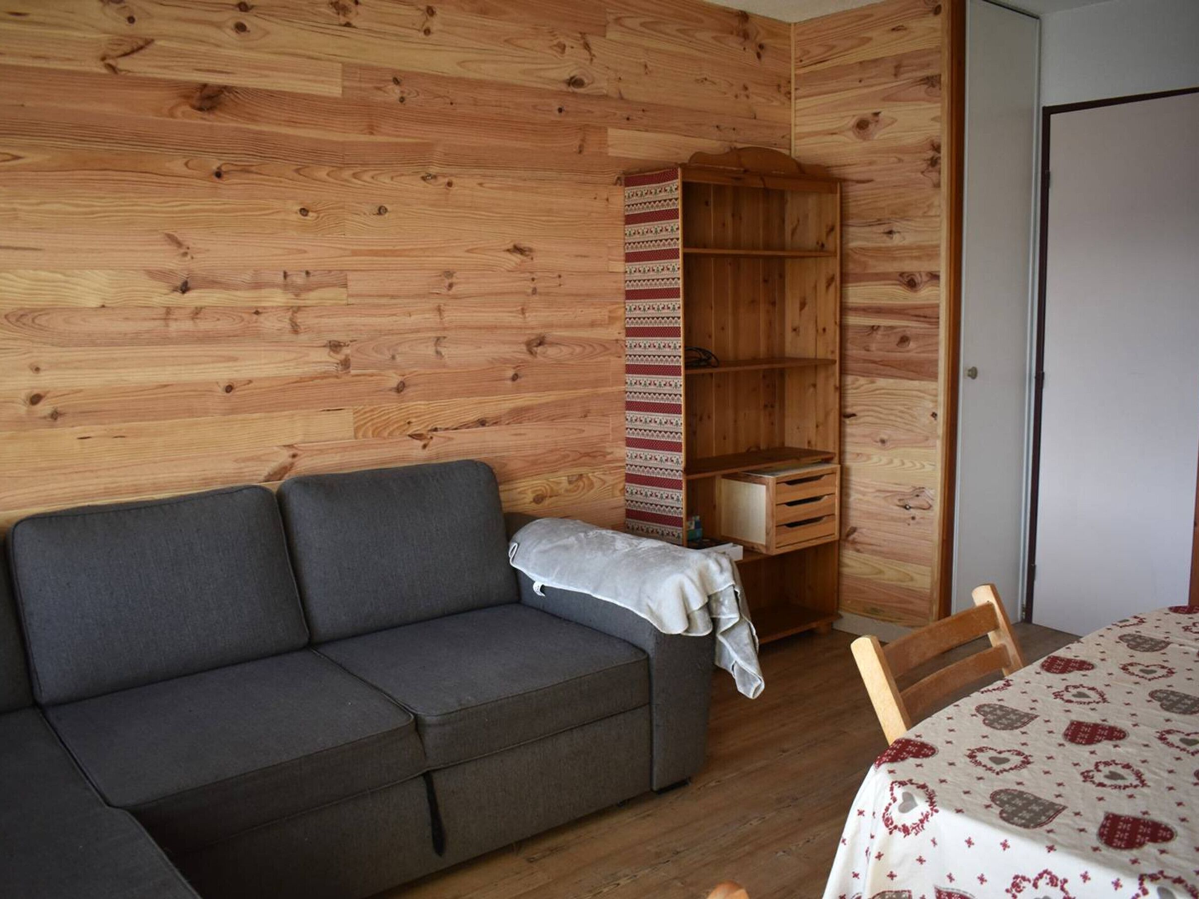 A room with a grey sofa, a wooden bookshelf, and a table with a patterned tablecloth.