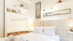 A hotel room with a bed, a bedside table with a lamp, a wall-mounted mirror, and shelves with books and a plant. - Provençal Mazet with Pool, Pets Welcome, Secure Parking (Le Lavandou)