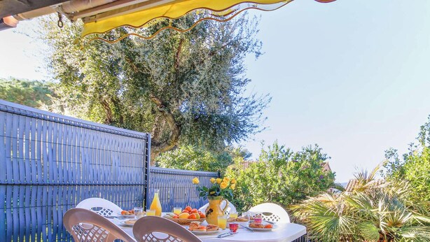 An outdoor dining area with a table set for two, surrounded by lush greenery and a blue fence. - Provençal Mazet with Pool, Pets Welcome, Secure Parking (Le Lavandou)