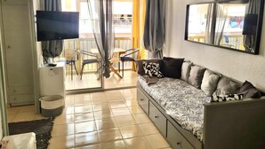 A compact living space with a sofa, a TV, a dining area with chairs, and a balcony with curtains. - Beautiful beachfront apartment with air conditioning, pool, and parking in La Pineda (La Pineda)