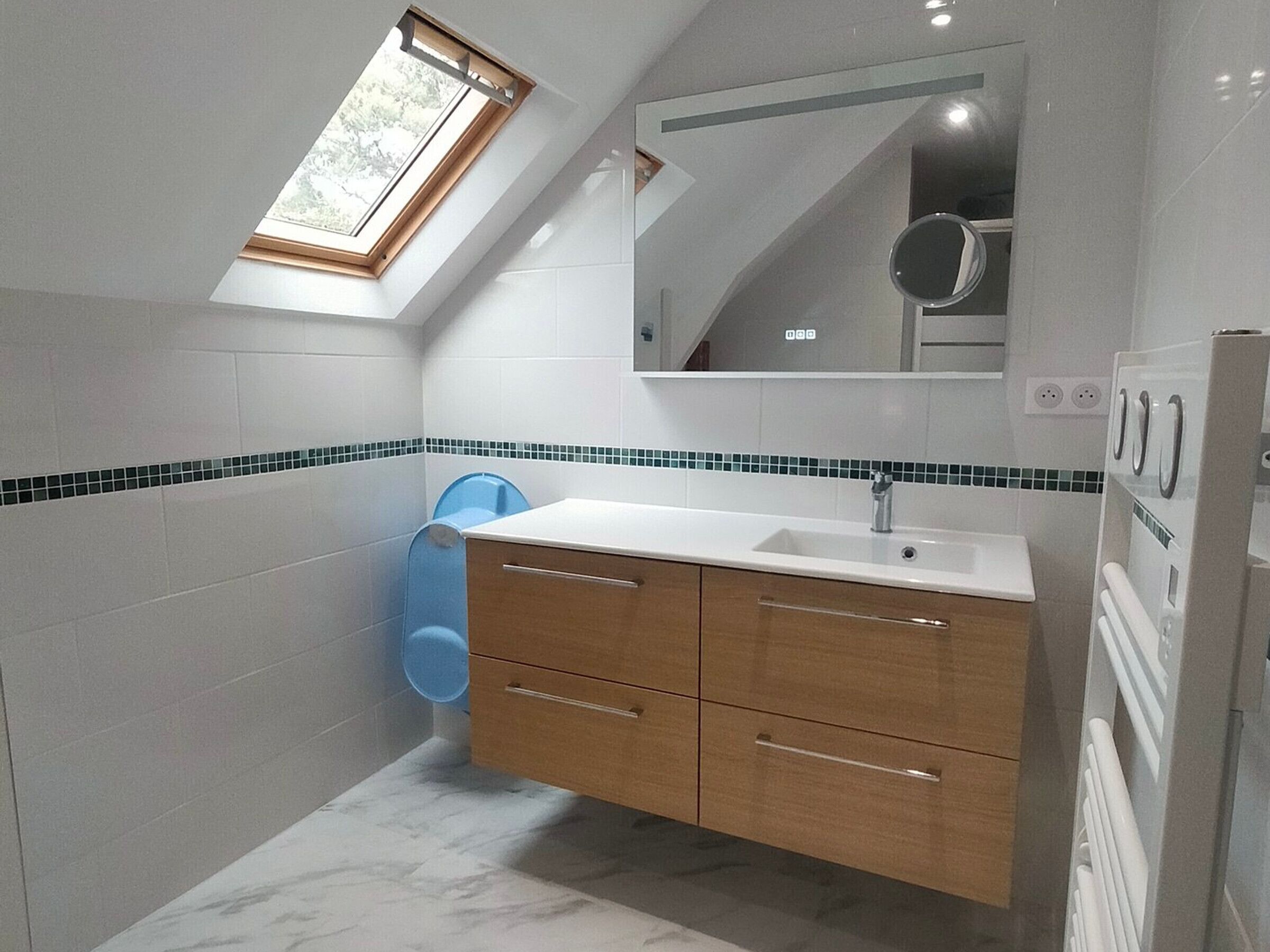 Ein modernes Badezimmer mit Dachfenster, weißen Fliesen, einer blauen Toilette und einem hölzernen Waschtisch mit Schubladen.