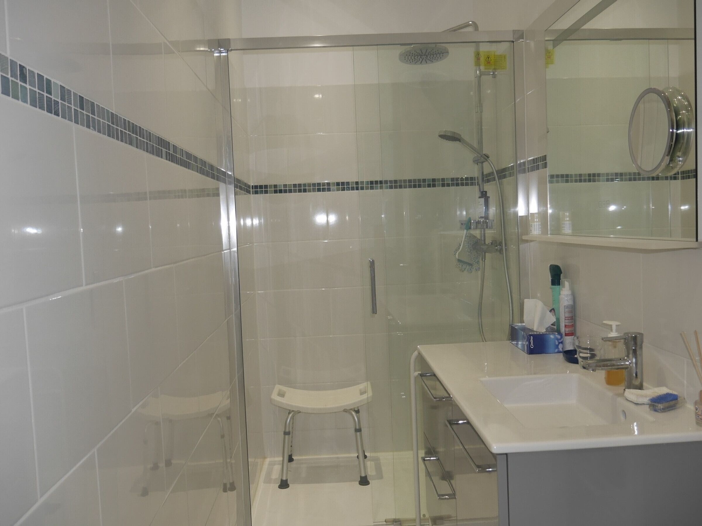 Une salle de bain moderne avec une douche à parois de verre, un lavabo blanc avec un miroir au-dessus, et un petit tabouret.