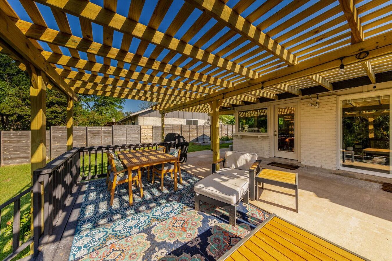 Ein überdachter Patio-Bereich mit einer Holz-Pergola, Gartenmöbeln und einem gemusterten Teppich.