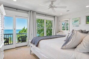 A bedroom with a bed, a ceiling fan, a view of the ocean through large windows, and a balcony with chairs. - Beachfront Haven | Grill • Hot Tub • Sunset Views! (South Haven)