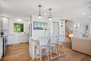 A modern kitchen with white cabinets, a central island with chairs, and a painting of sailboats. - Beachfront Haven | Grill • Hot Tub • Sunset Views! (South Haven)