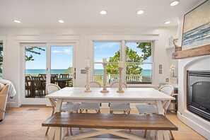 A dining area with a table and chairs, a fireplace, and large windows with a view of the ocean. - Beachfront Haven | Grill • Hot Tub • Sunset Views! (South Haven)