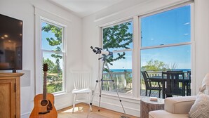 Ett rum med en gitarr i trä, ett teleskop, en vit stol och utsikt över naturen genom stora fönster.