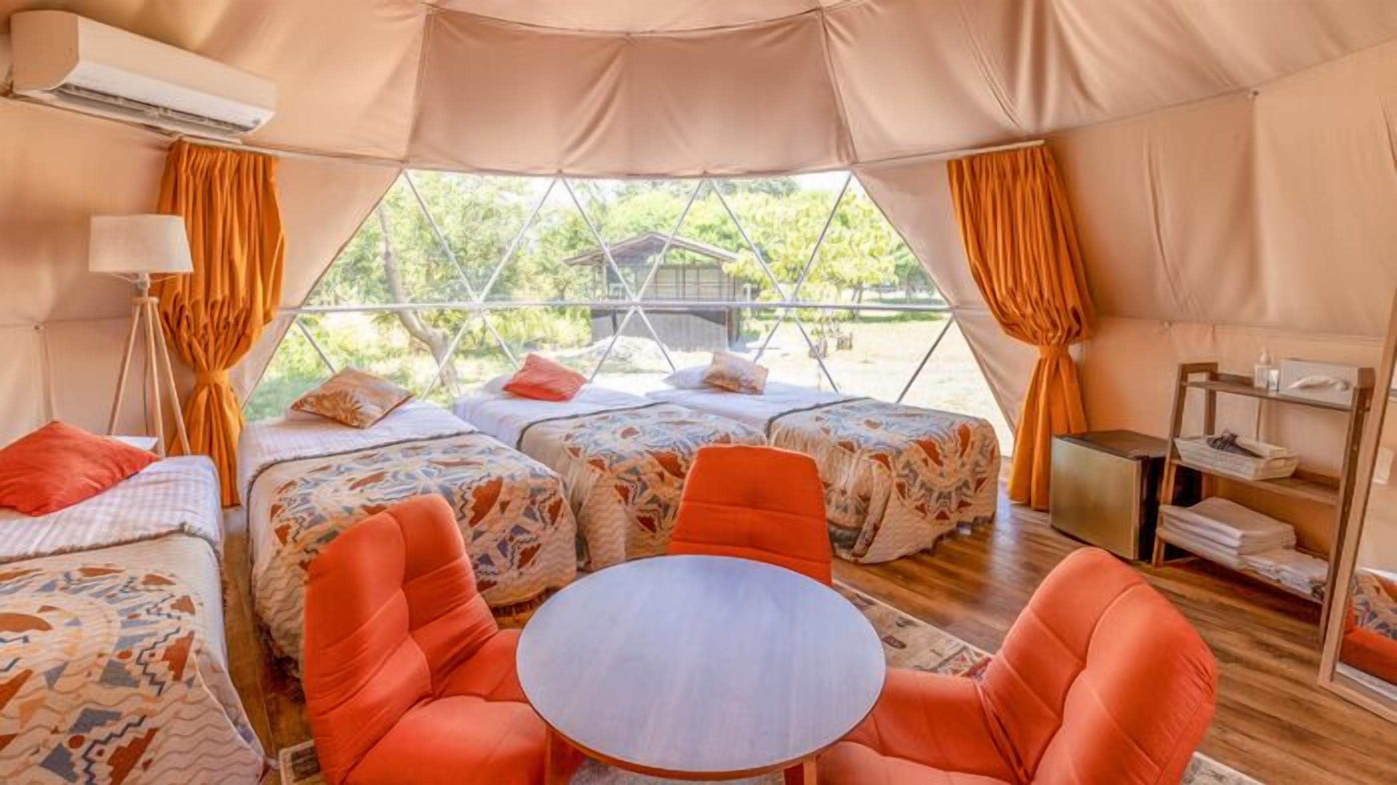 A tented accommodation with two beds, a round table, and orange chairs.