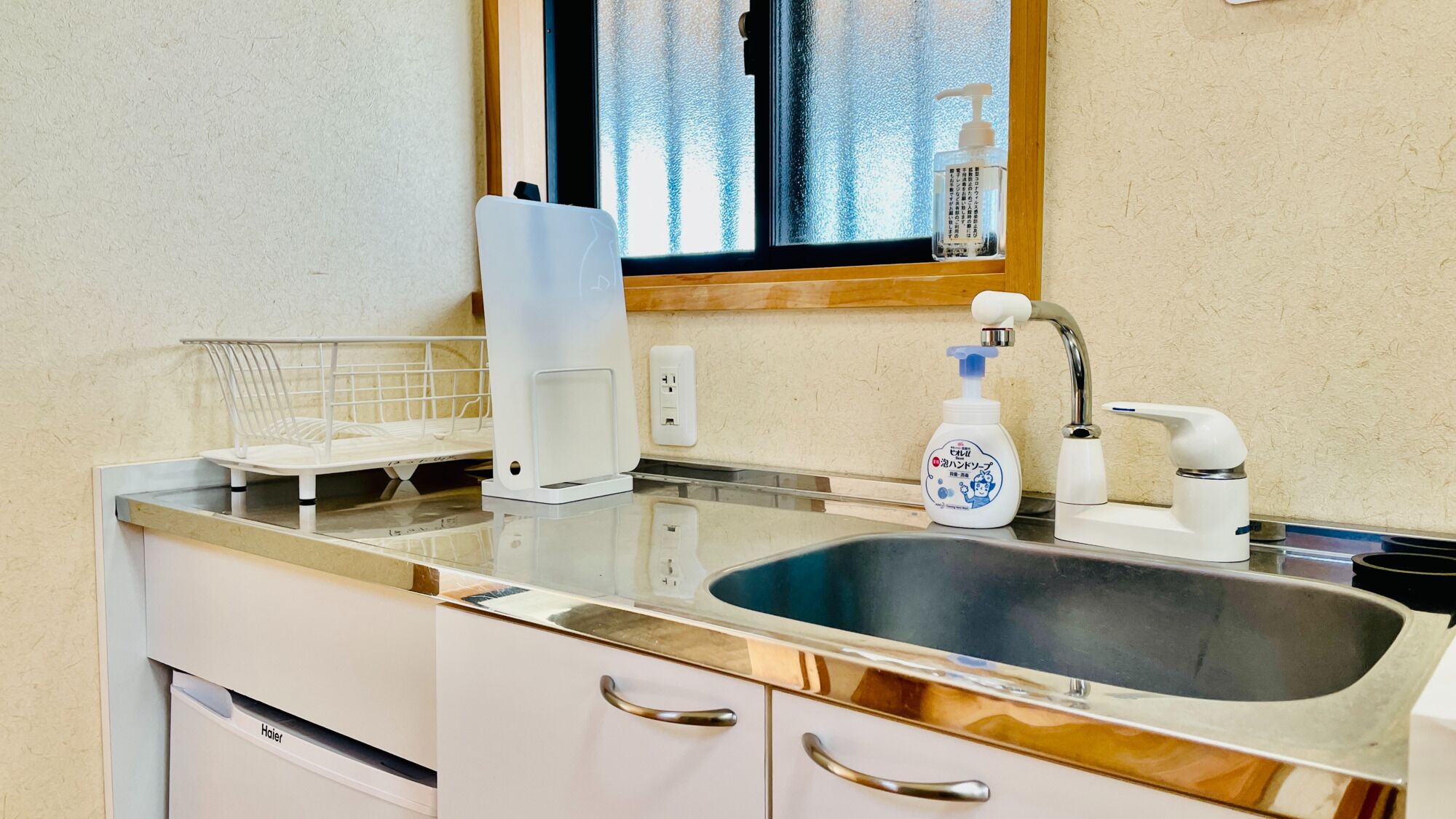 A kitchen with a stainless steel sink, a white countertop, a window with a view, and a bottle of hand soap.