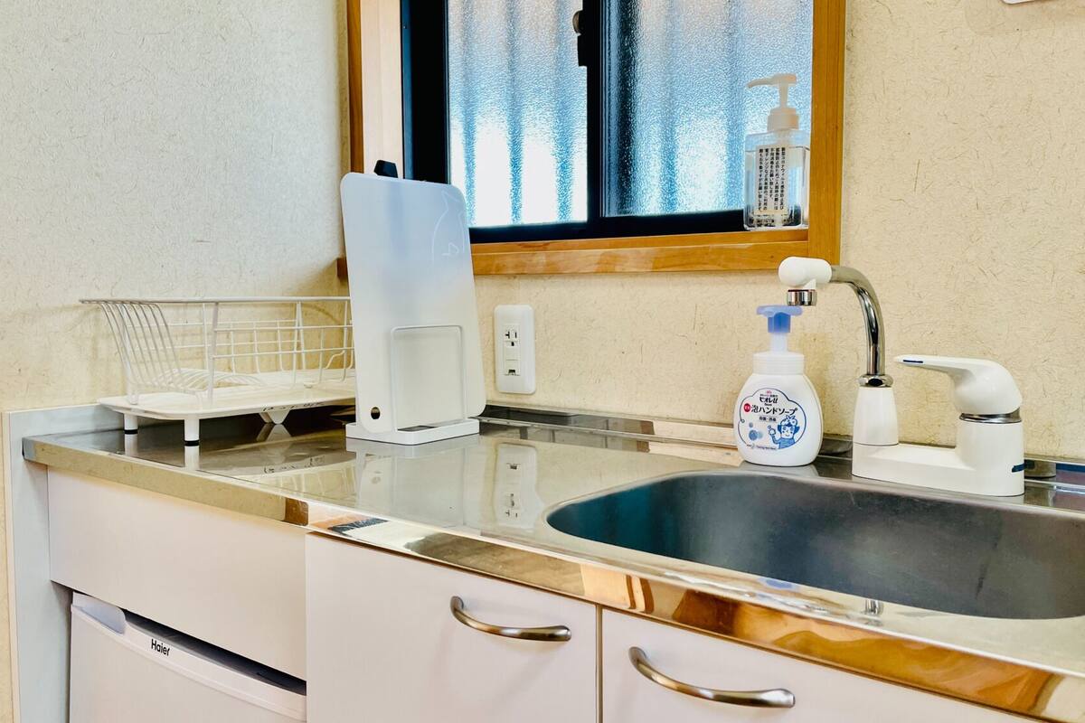 Una cocina con un lavamanos de acero inoxidable, una encimera blanca, una ventana con vistas y una botella de jabón para manos.