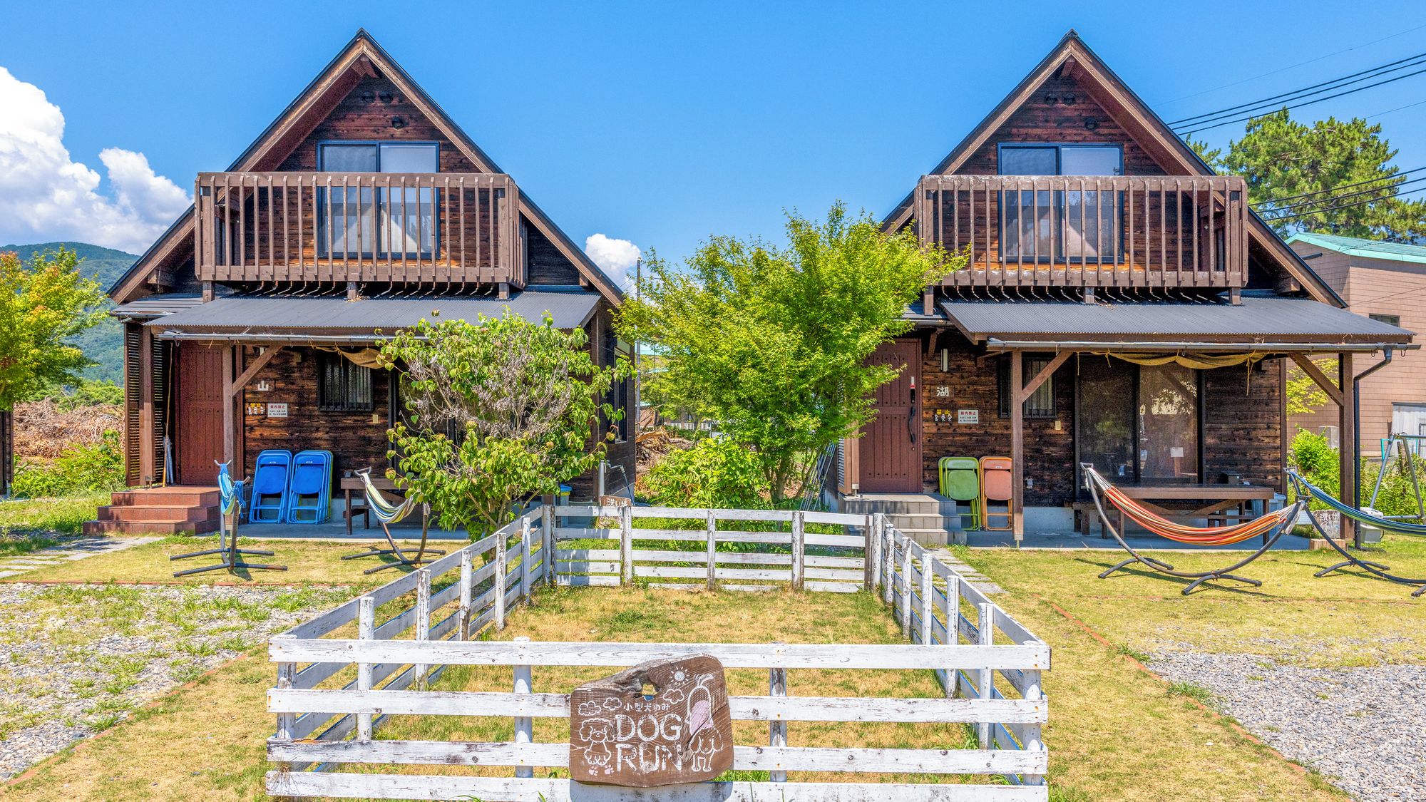 Dos cabañas de madera con balcones, un área cercada y una hamaca.
