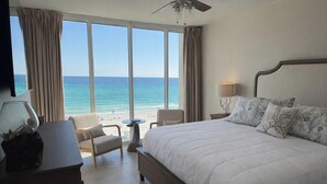 Quarto de hotel com uma cama grande, uma escrivaninha, uma cadeira, um abajur e vista para a praia e oceano.