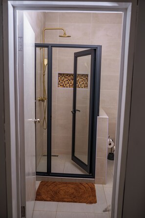 Une douche fermĂ©e par des parois de verre, dotĂ©e dâun pommeau de douche dorĂ©, un tapis marron au sol et un mur carrelĂ© avec une petite section en mosaĂŻque.