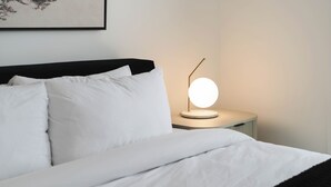 A neatly made bed with white linens and a black headboard. A round, gold-framed table lamp with a white shade is on a bedside table. - Veldt Haus (Revelstoke)