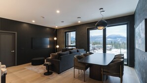 A modern living room with a dark wood panel wall, a large flat-screen TV, a dark sofa, a dining table with chairs, and large windows offering a view of a snowy landscape. - Veldt Haus (Revelstoke)