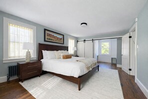 Une chambre à coucher avec un grand lit, une table de chevet, une commode, un radiateur et une fenêtre avec des rideaux.