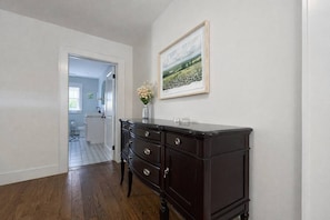 Ein Flur mit Holzboden, einer dunklen, schubladigen Kommode sowie einem gerahmten Landschaftsbild an der Wand und einer Tür zu einem weiteren Raum.