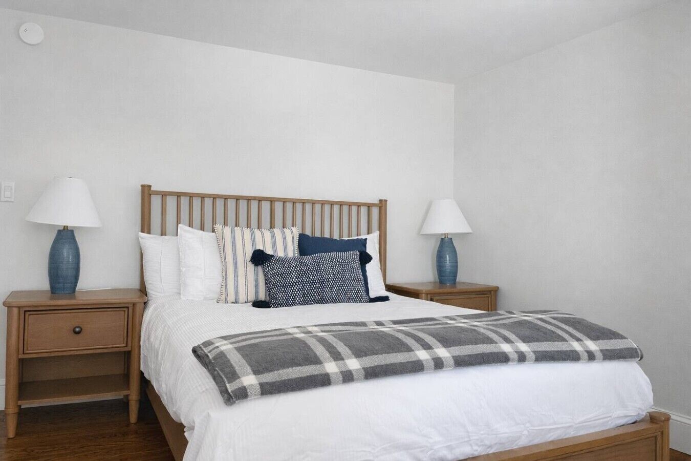 Een slaapkamer met een houten bedframe, wit beddengoed, twee nachtkastjes met lampen en een geruit plaid.