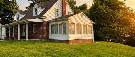Charming two-story home featuring classic brick and white siding, a covered front porch, a sunroom, and an expansive grassy yard framed by mature trees.