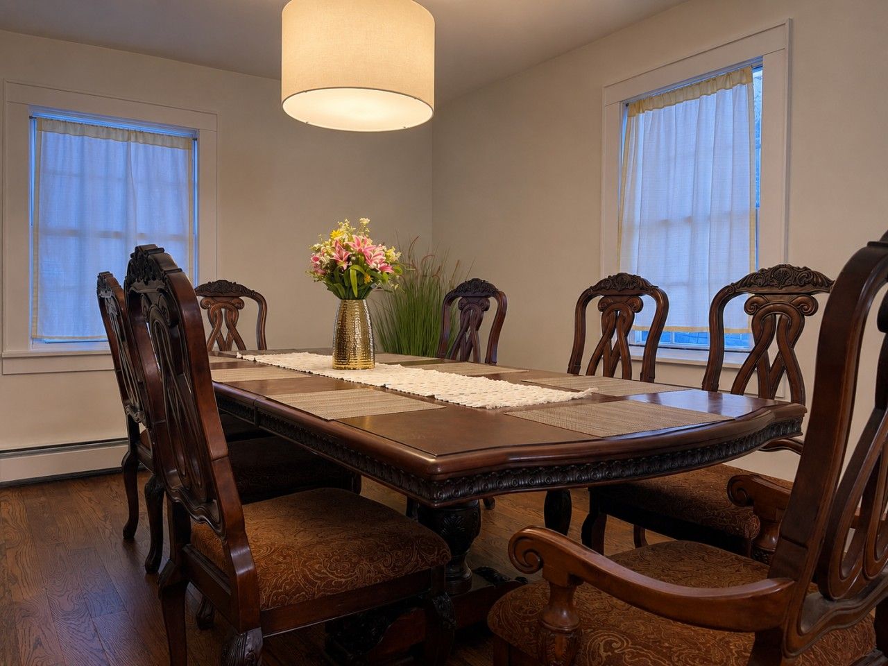A dining room with a rectangular table, six wooden chairs, a vase with flowers, and two windows with curtains.