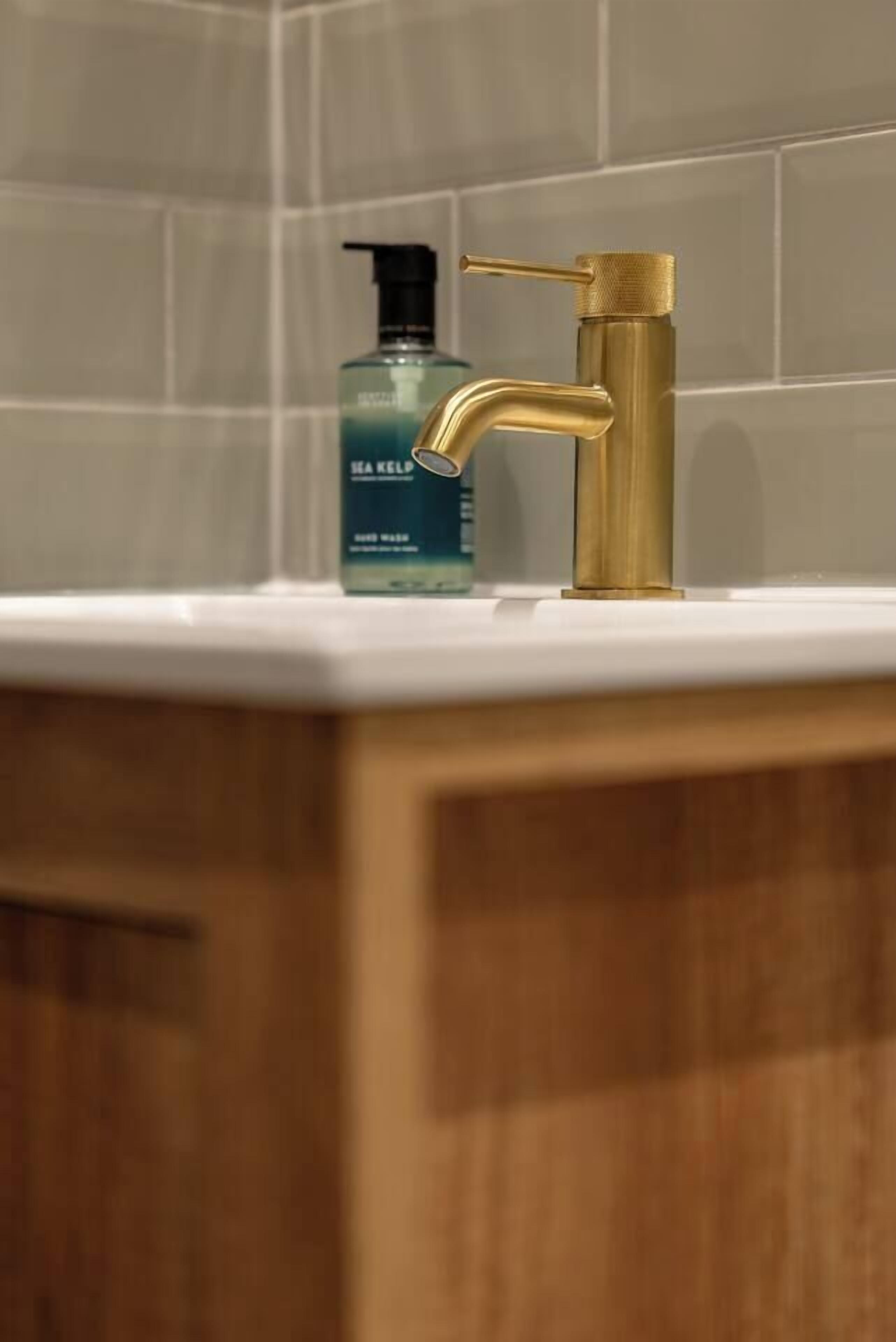 Une salle de bain avec un robinet doré, une bouteille bleue de produit à base d’algues marines et des carreaux blancs.