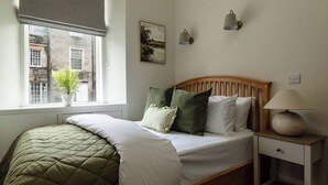 Ein Schlafzimmer mit Bett, Nachttisch, Lampe, Fenster mit Blick auf Gebäude und einer Topfpflanze.