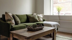 Ein grünes Sofa mit Kissen, ein Couchtisch aus Holz mit einem Korb und ein Fenster mit Blick auf ein Gebäude.