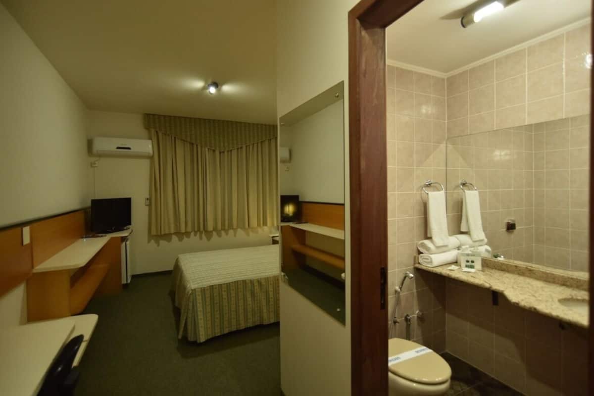 Una camera d'albergo con un letto, una scrivania con un computer, una televisione, un bagno con il water e uno spazio doccia.
