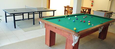 Una mesa de billar con bolas, una mesa de ping pong y una mesa de fútbolín en una sala de diseño sencillo.