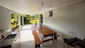 A kitchen with a sink, a wooden table, and a bench. There is a window with a view of a green landscape and a cow. - Pousada Praia Verde - Ilha Redonda | Palmitos-SC (Palmitos)