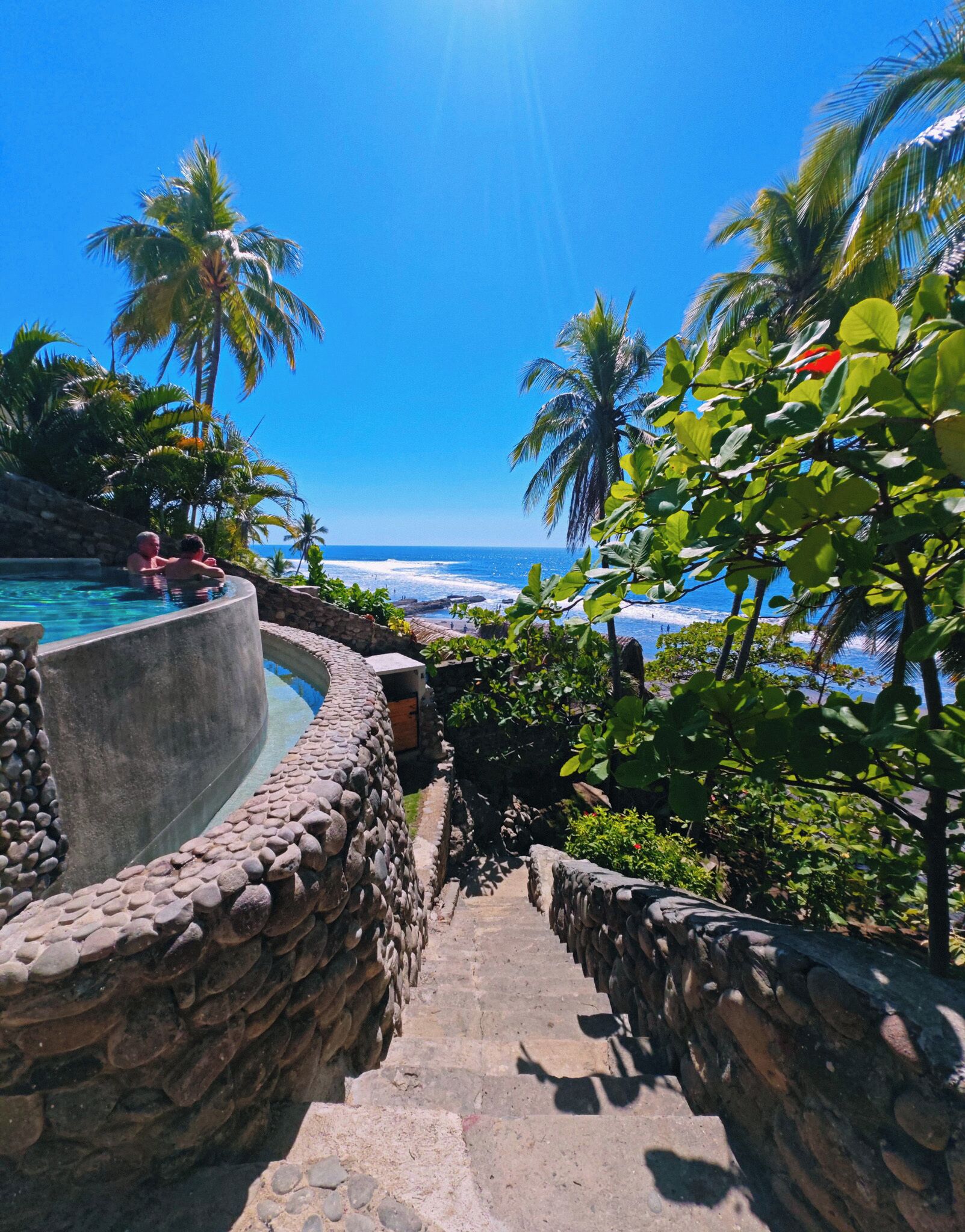 A stone staircase leading to a pool with a view of the ocean and palm trees.