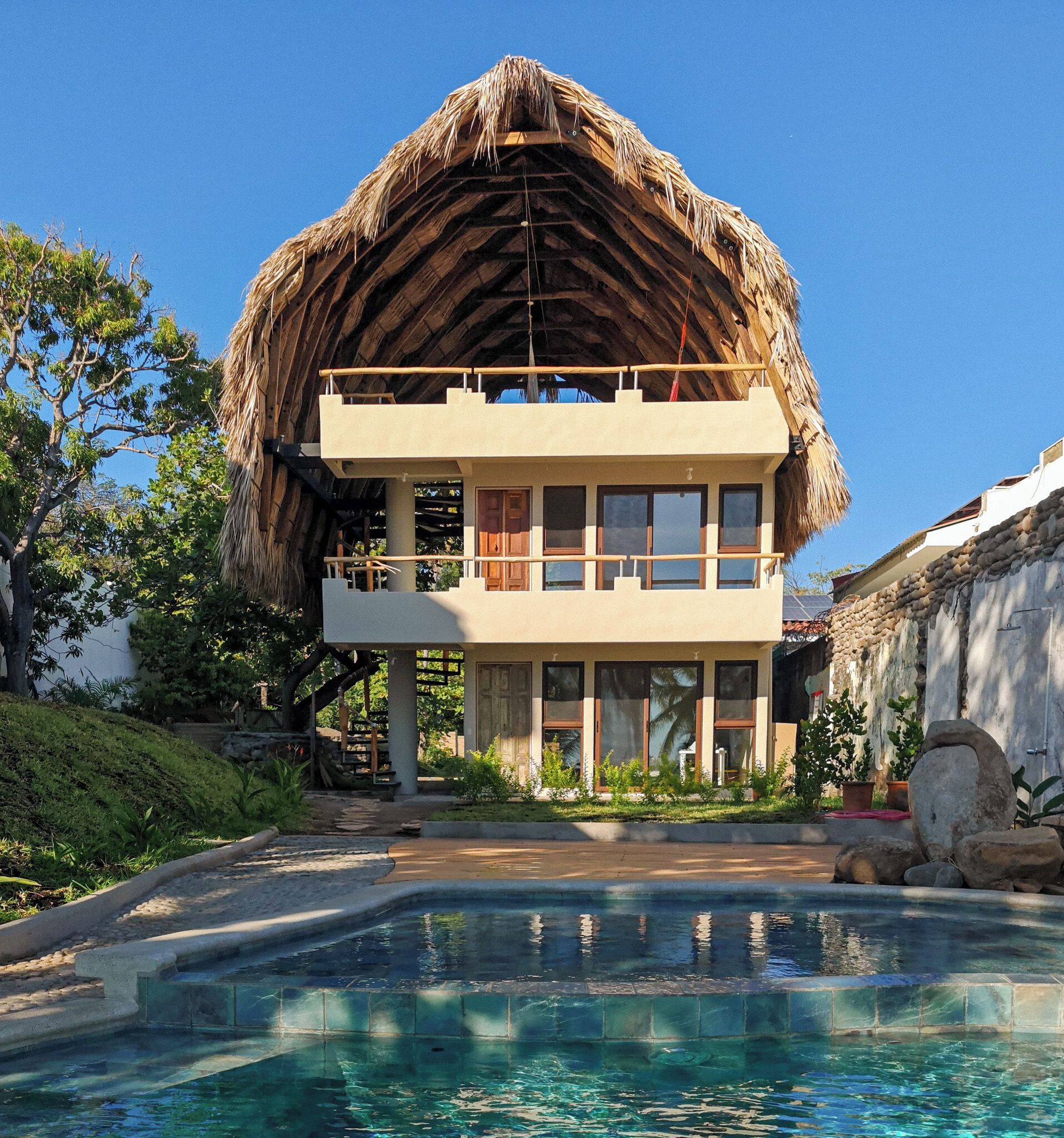 A two-story building with a thatched roof, a swimming pool, and a garden with tropical plants.