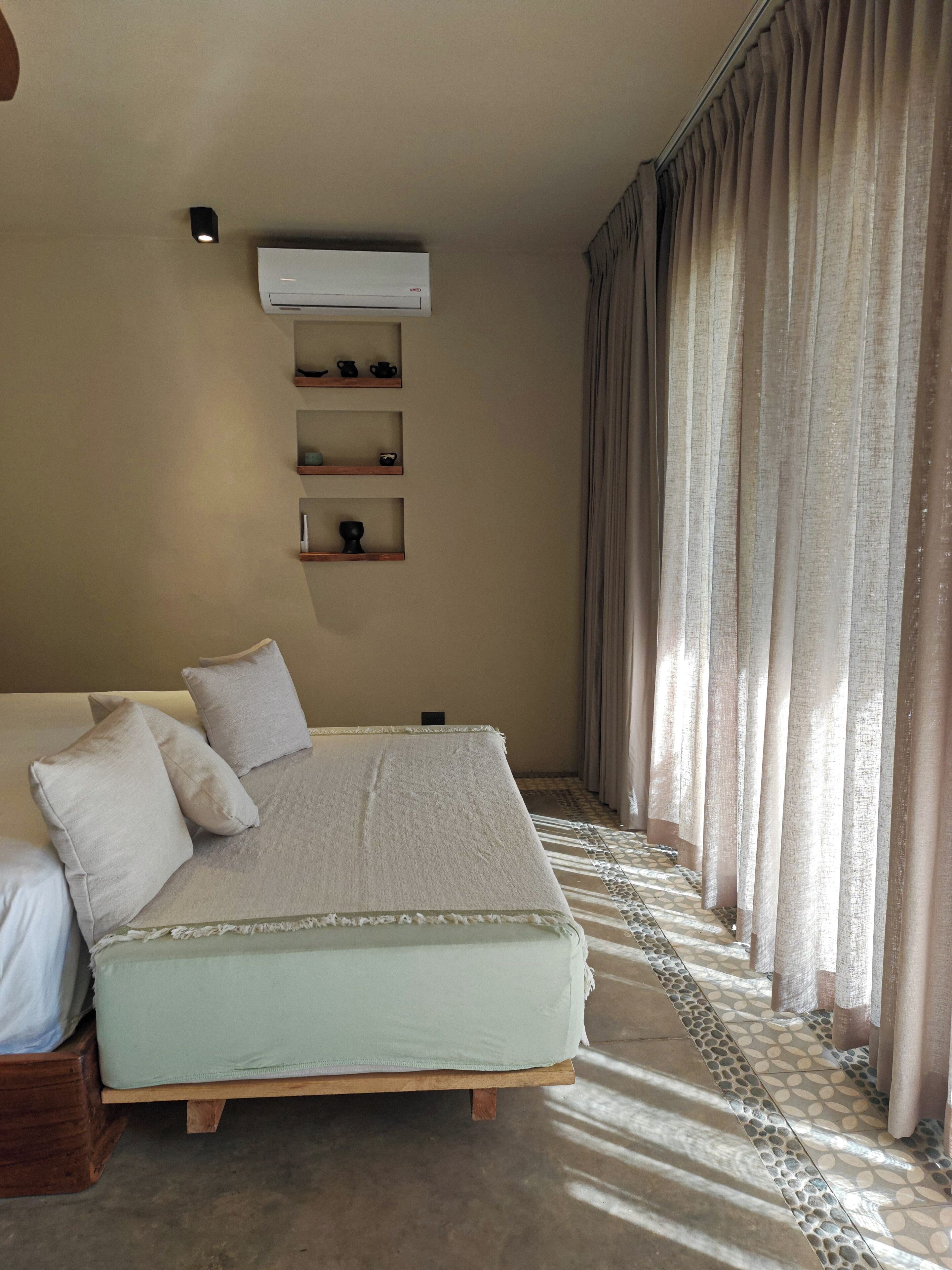 A bedroom with a bed, a wall-mounted shelf with items, and a window with curtains.