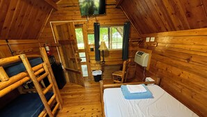A wooden cabin with bunk beds, a small table, a chair, a TV, and a window with curtains.