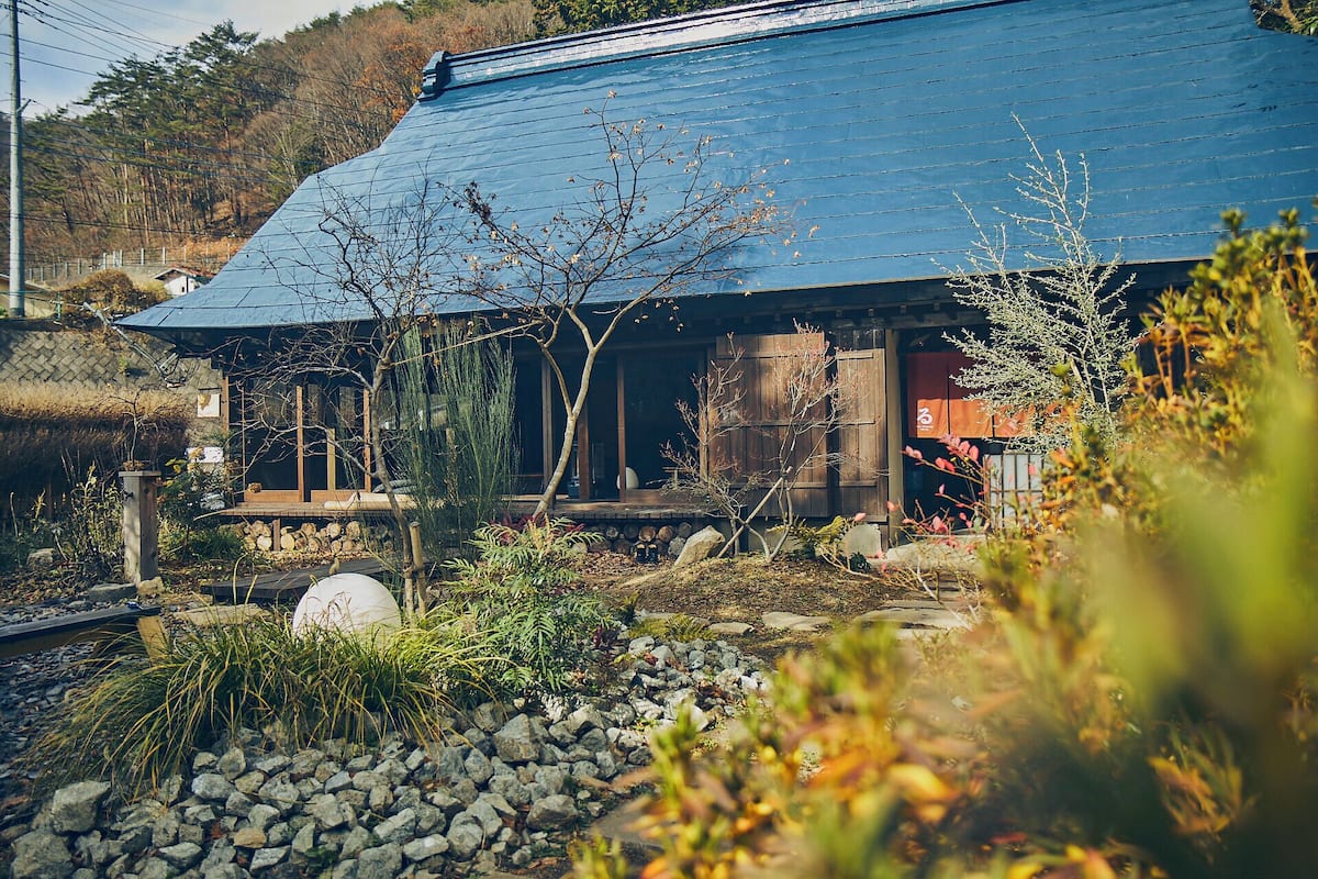 A traditional Japanese house with a sloped roof, wooden exterior, and a garden with rocks and sparse vegetation.